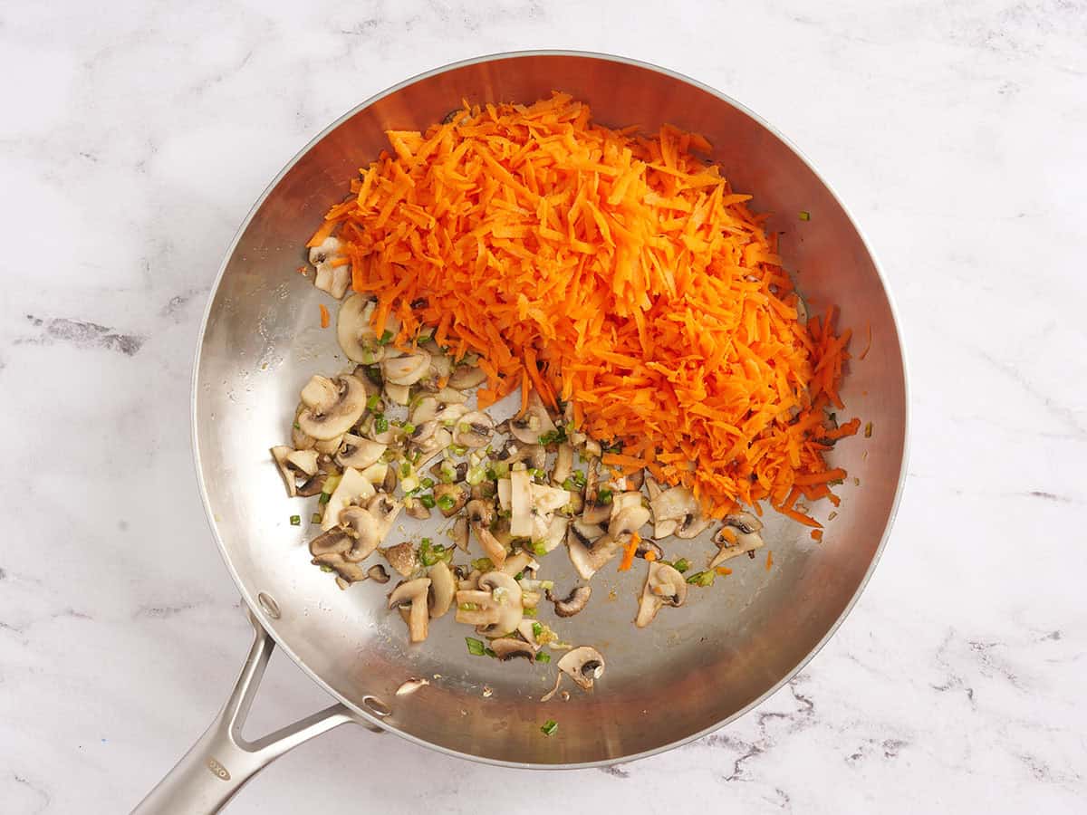 Shredded carrots added to a skillet of sauteed mushrooms.