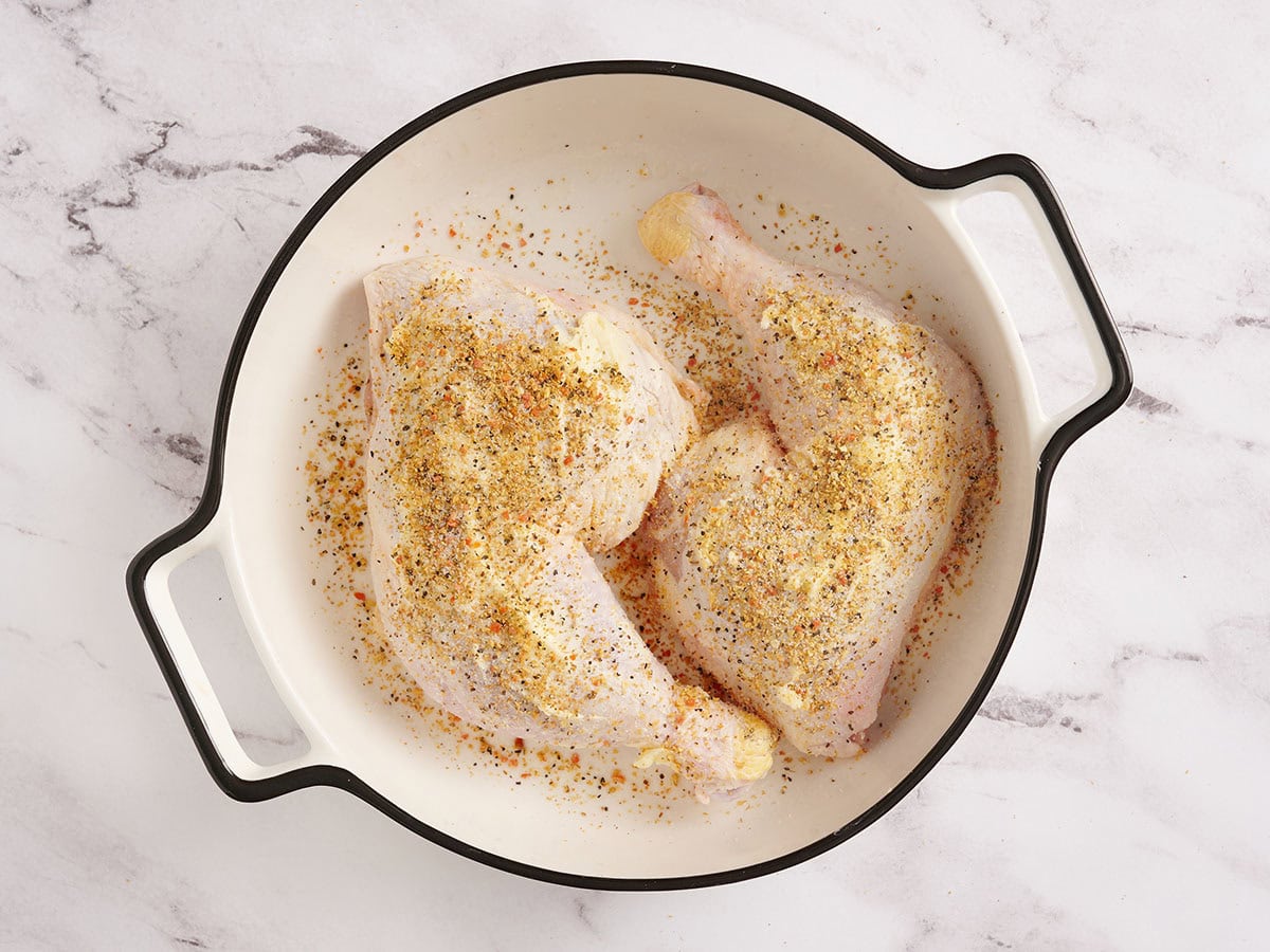 Two raw chicken legs in a baking dish covered in butter and seasonings.