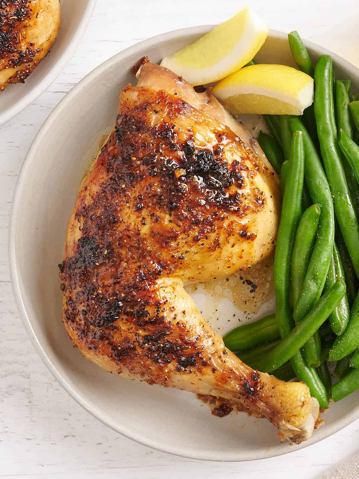 Slow and low heat is the key to tender and juicy oven roasted chicken legs. This simple recipe makes an easy main dish for any night of the week. Budetbytes.com Overhead view of a baked chicken leg on a plate with green beans and lemon wedges.