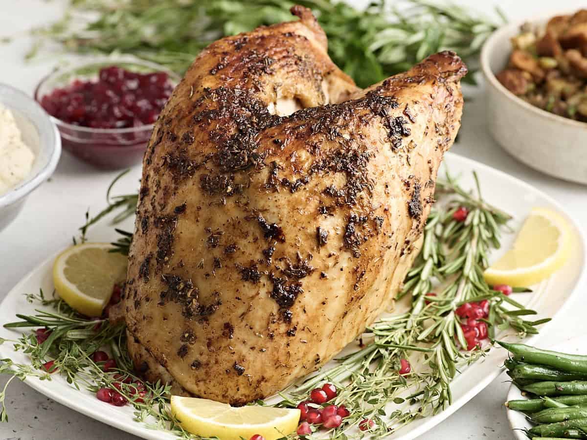 Side view of a cooked turkey breast on a white platter garnished with fresh herbs and lemon.