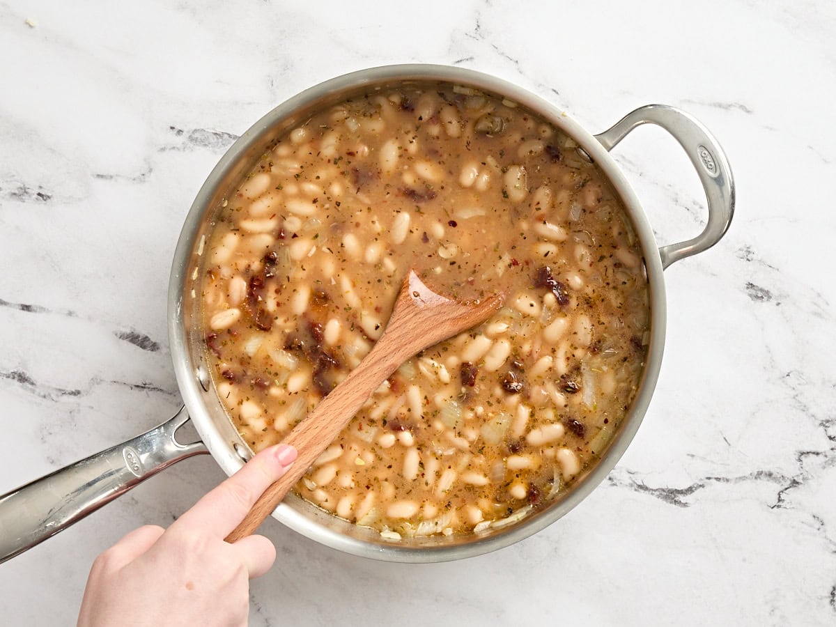 A wooden spoon mashing some white beans in a skillet.