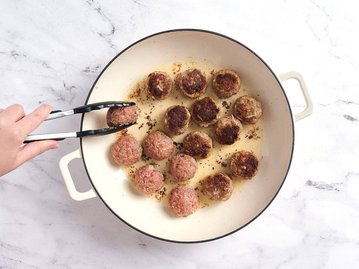 Swedish meatballs cooking in a pot, with tongs flipping them over.