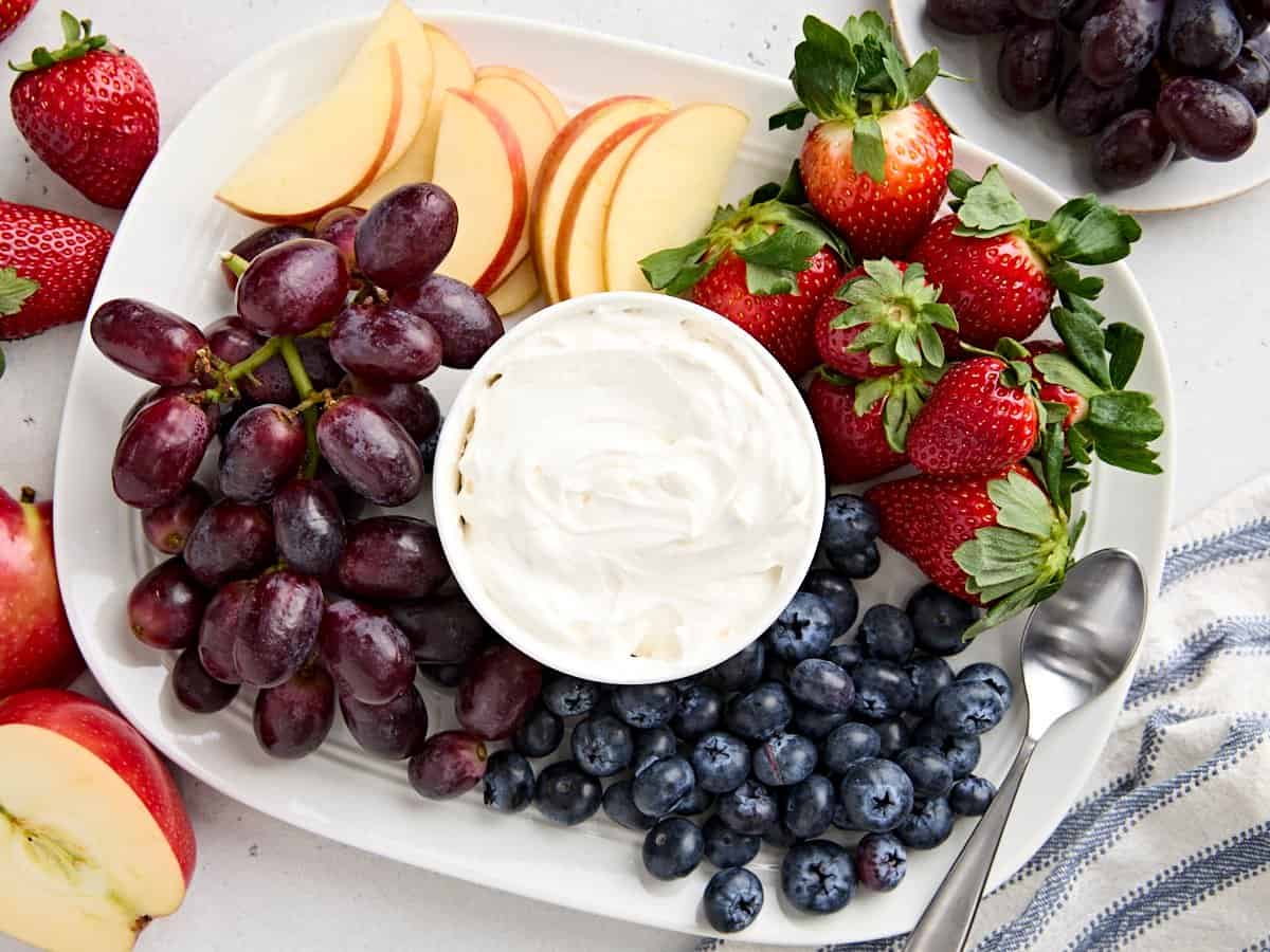 Overhead view of a bowl of homemade fruit dip surrounded by grapes, apple slices, blueberries, and strawberries.