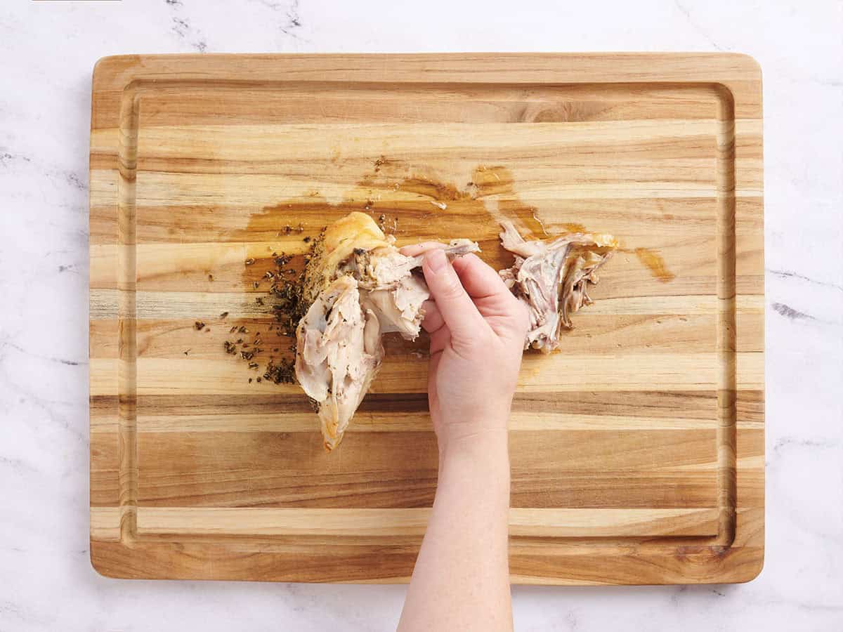 Hands removing the bone from cooked chicken breasts on a wooden cutting board.