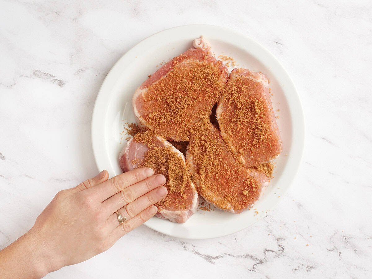 Four pork chops on a plate being rubbed with a brown sugar seasoning rub.