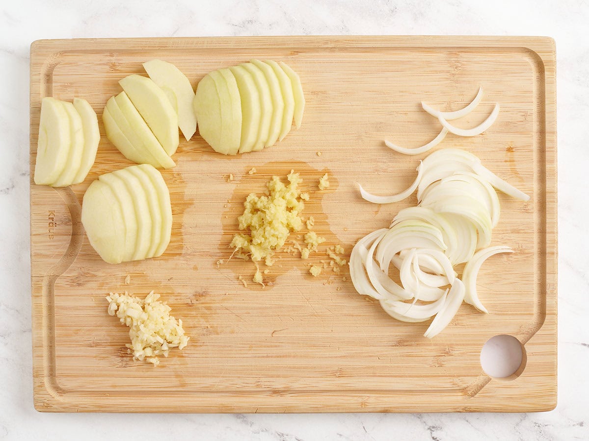 A peeled and sliced green apple, grated ginger, minced garlic, and sliced onion on a wooden chopping board.
