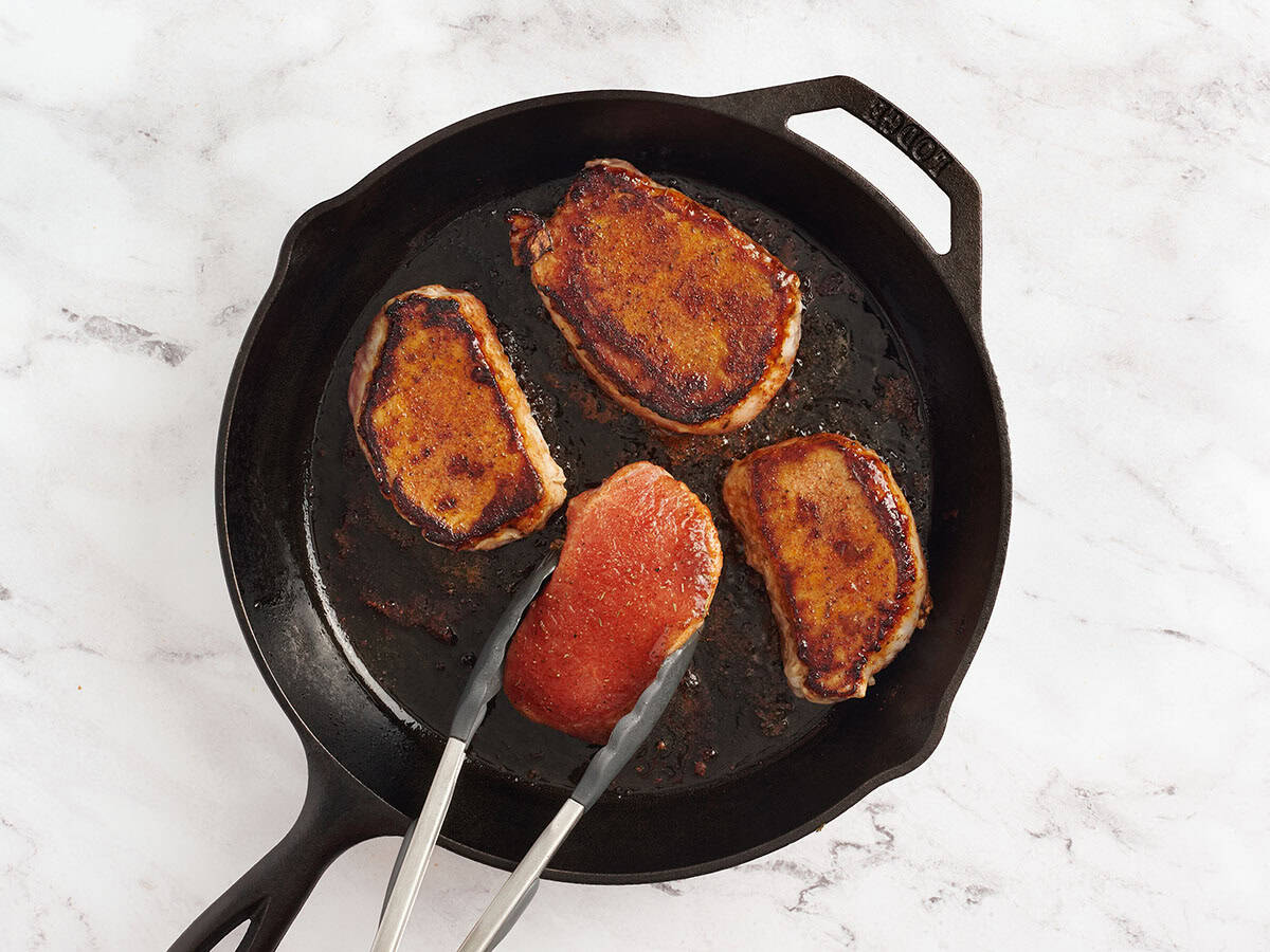 Kitchen tongs flipping over seasoned pork chops in a skillet.