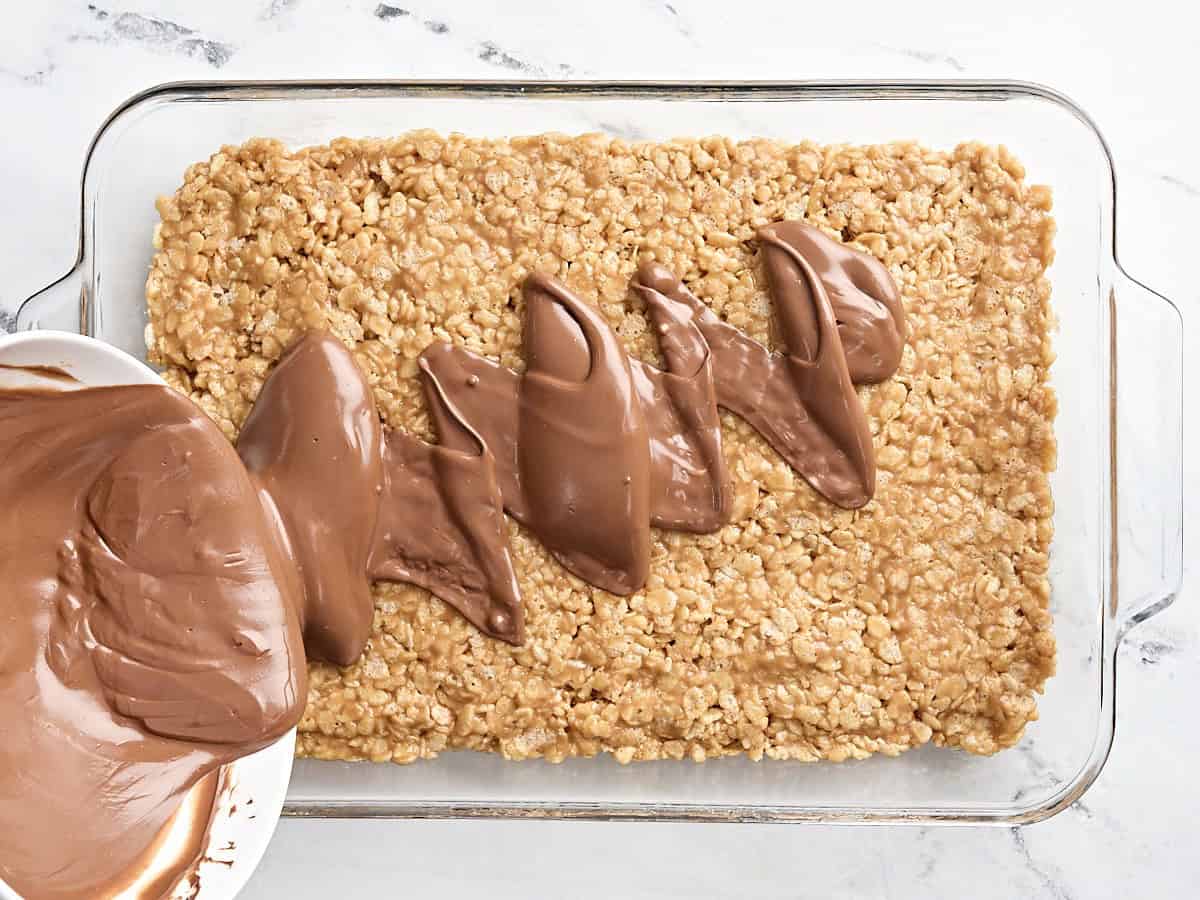 A bowl pouring melted chocolate over the rice krispie dough for Scotcharoos in a glass baking dish.