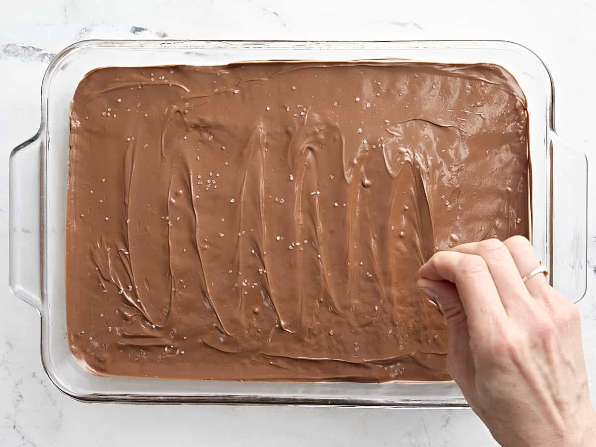 A hand sprinling sea salt over Scotcharoos in a glass baking dish.