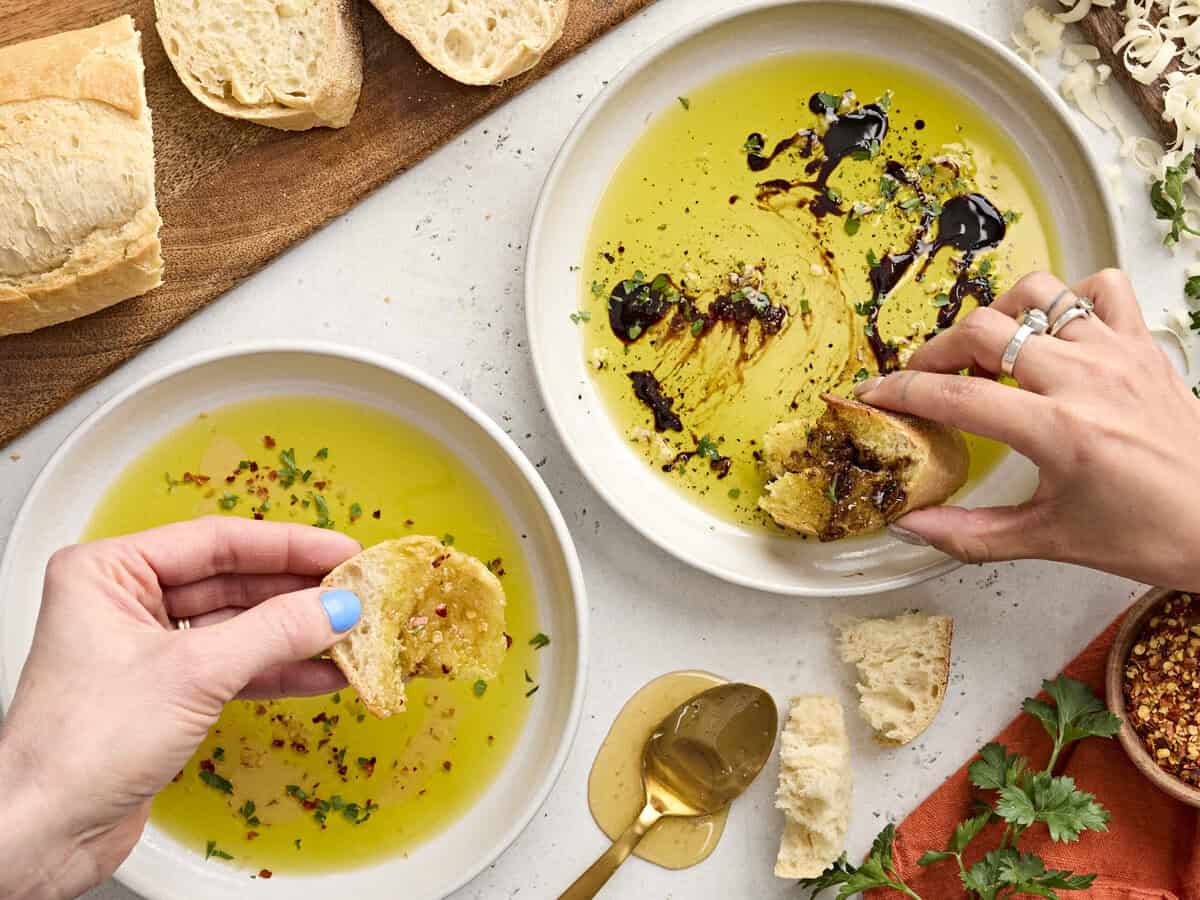 Two bowls of olive oil dip with hands dipping bread. 