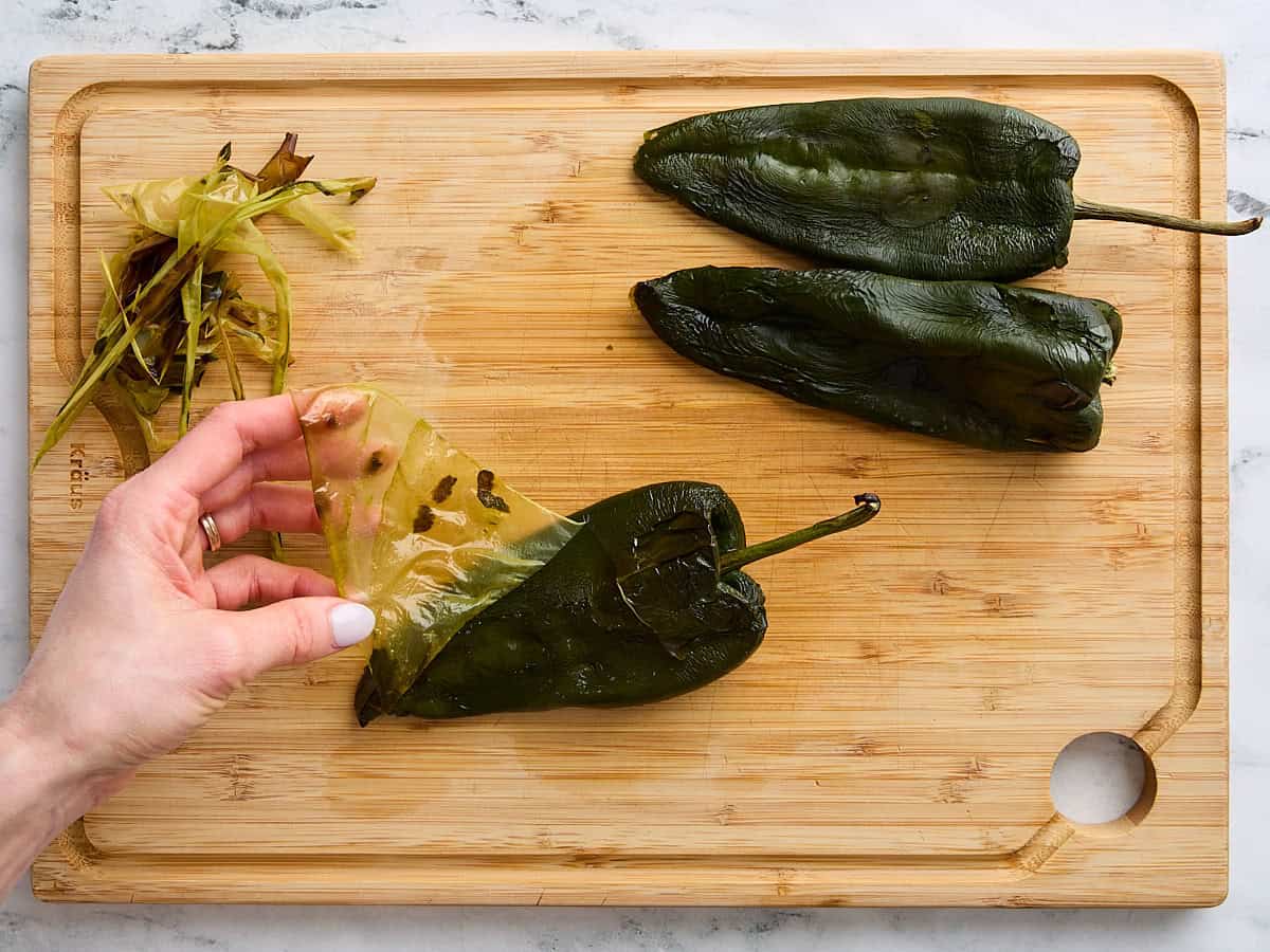 Three roasted poblano peppers on a wooden cutting board, with a hand peeling the skin from one.