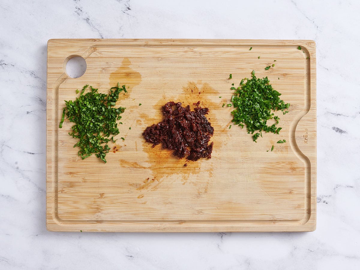 Minced fresh herbs and sun dried tomatoes on a wooden cutting board.