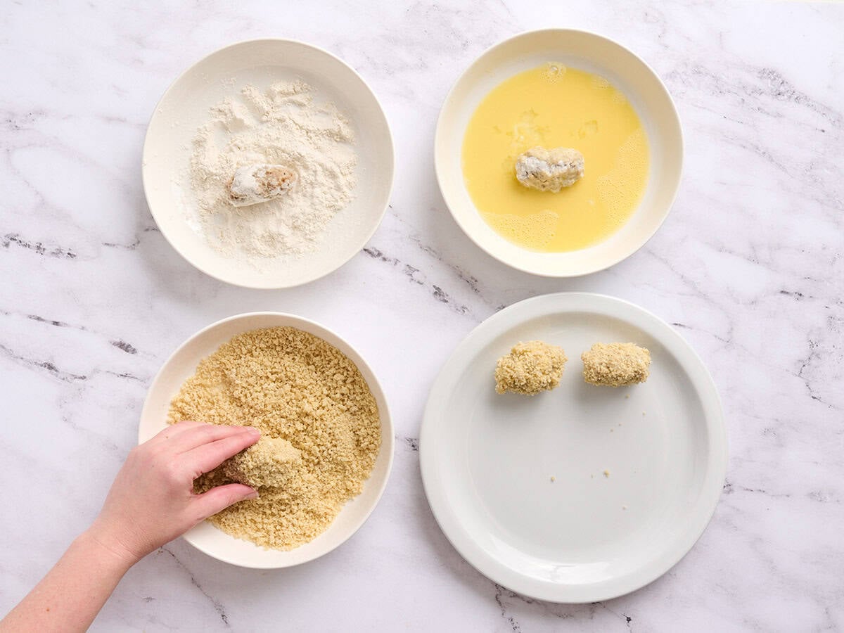 Four bowls, one with flour, one with beaten egg, one with breadcrumbs and one with breaded chicken croquettes.