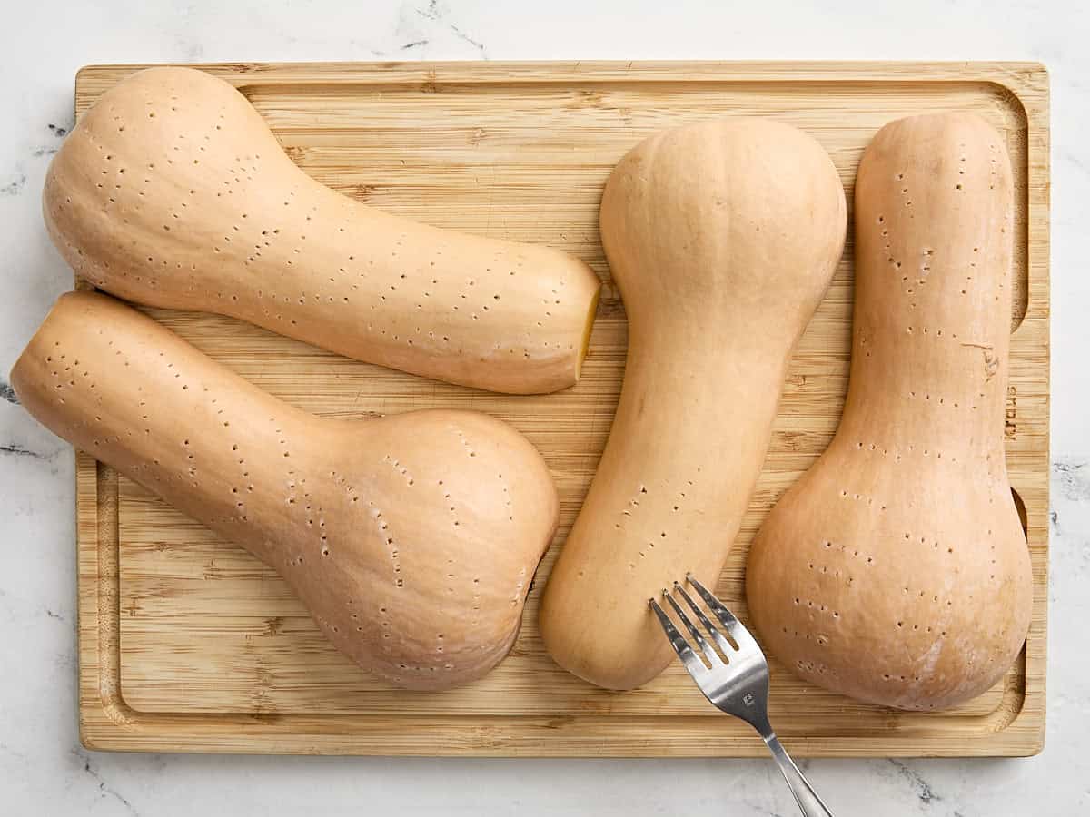 A fork poking hole in the skin of 4 halved butternut squash on a wooden chopping board.