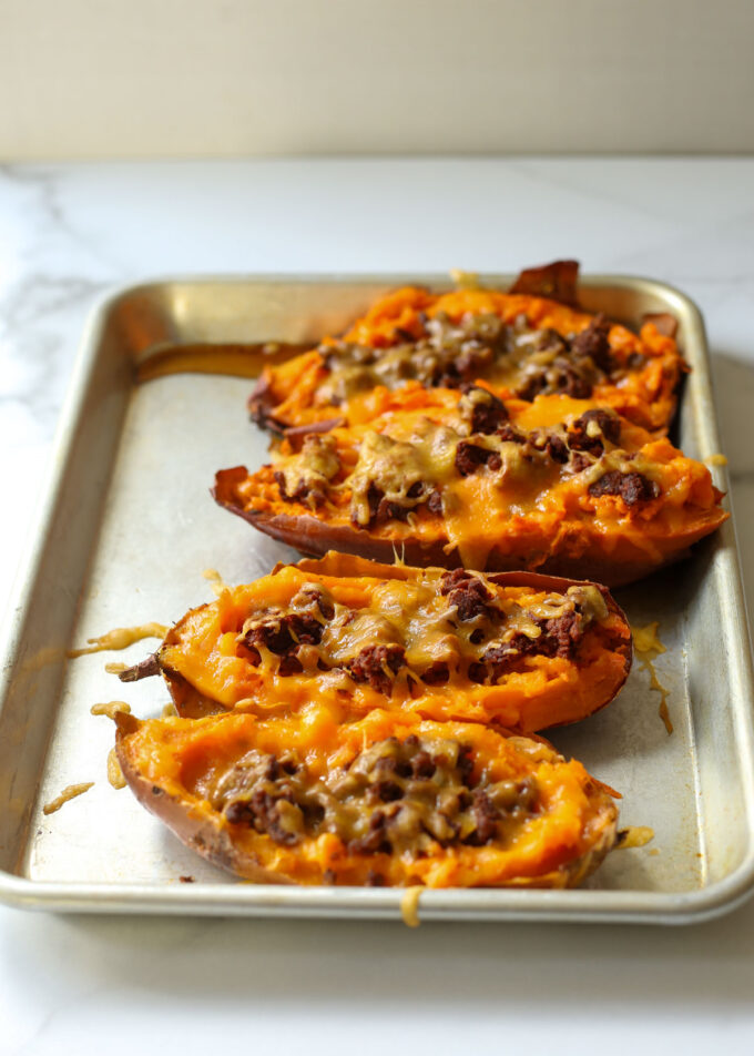 A metal rimmed baking pan with halved baked sweet potatoes, meat/vegan meat filling, and cheese, baked in the oven until warm.