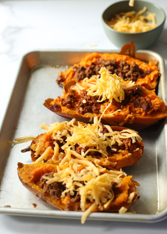 A metal rimmed baking pan with halved baked sweet potatoes, meat/vegan meat filling, and cheese, ready to bake.