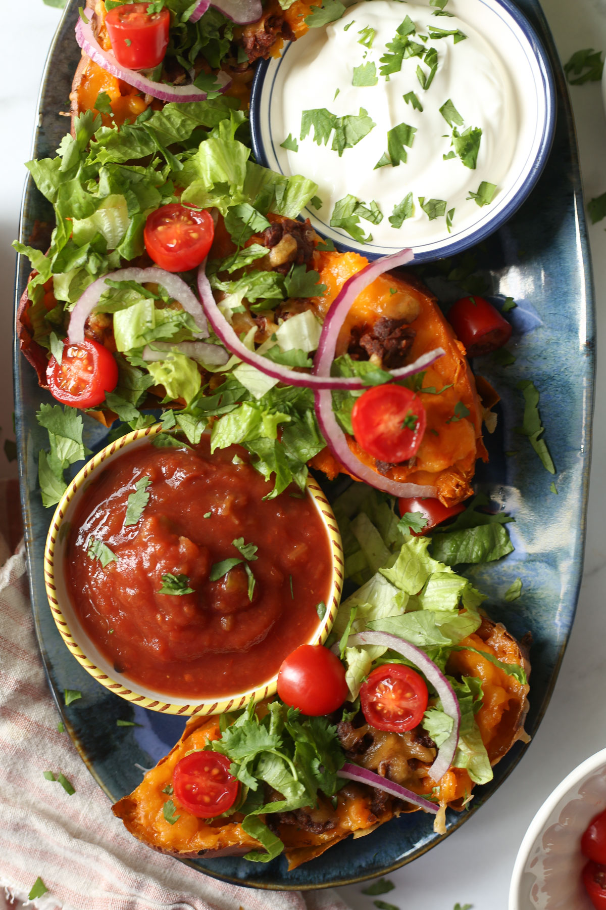 A top view of a blue platter with taco sweet potatoes, salsa, and sour cream. A pink and white striped napkin and various toppings are in the background.