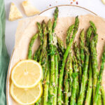 A top view of a white plate lined with parchment paper and parmesan roasted asparagus on top with lemon as garnish.