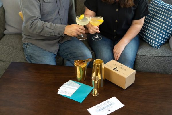 box of barware with 2 cocktails being held by hands, surrounded by gift certificates for a cocktail cooking class